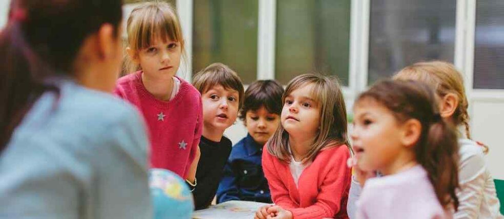 Eine Gruppe von Vorschulkindern steht an einem Tisch und hören interessiert den Erklärungen der Lehrerin zu.