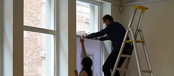 Ein Mann auf einer Trittleiter und eine Frau hängen eine Zeichnung in ein Fenster. 
