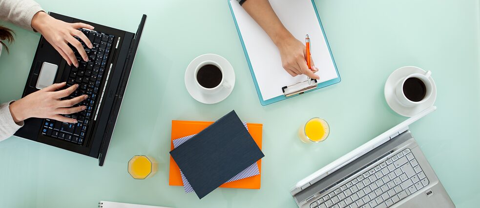 Draufsicht auf einen Tisch, auf dem sich Tablet, Laptop, Schreibblock, Kaffeetassen und Gläser mit Orangensaft befinden.
