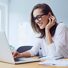 Eine Frau arbeitet in einem Büro am Laptop. Sie tippt auf der Tastatur und lächelt. Im Hintergrund ist eine Pinnwand und ein Flipchart, beides ist unscharf.