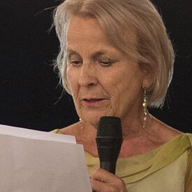 A woman with blonde hair is holding a microphone in her right hand. In her left hand she is holding papers with her speech, which she is reading. She is wearing an elegant green dress. The sky is dark and behind her there are fairy lights hanging.