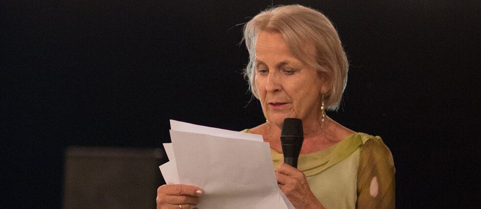 A woman with blonde hair is holding a microphone in her right hand. In her left hand she is holding papers with her speech, which she is reading. She is wearing an elegant green dress. The sky is dark and behind her there are fairy lights hanging.