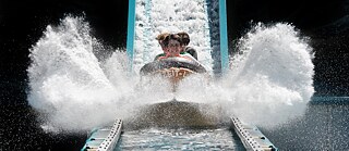 Frontal view of the vehicle of a log flume in which several people are sitting; water splashes very strongly on both sides