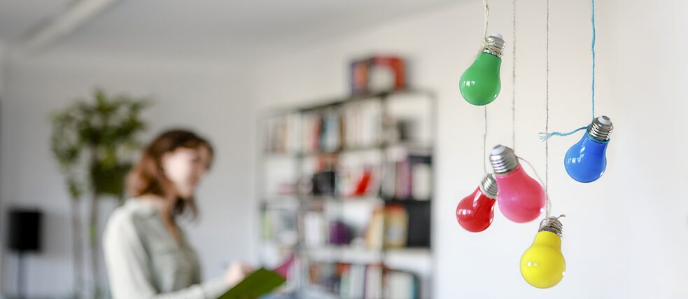 Colourful light bulbs dangle from the ceiling. A young woman with a book in her hands stands in the blurred background. 