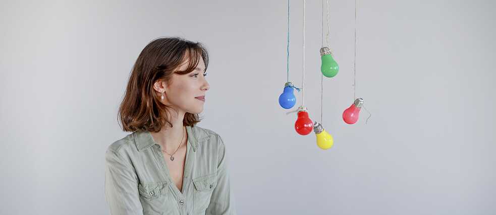 Eine junge Frau betrachtet bunte Glühbirnen, die in der Luft hängen. → A young woman is looking at colourful light bulbs dangling from a ceiling.