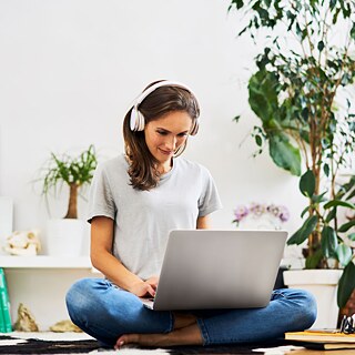 Eine Frau mit Kopfhörern sitzt zu Hause auf dem Boden und arbeitet lächelnd mit dem Laptop. Neben ihr stehen Zimmerpflanzen.