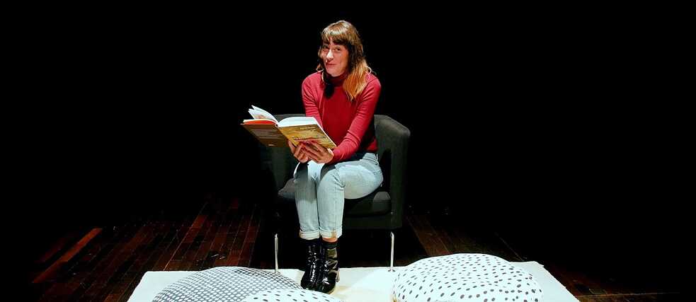 Deborah Weber lit un livre assise sur un fauteuil. Des coussins sont posés sur le sol devant elle.