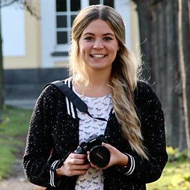 Rahel Klein hält eine Kamera in die Hand, hinter ihr ist ein gelbes Gebäude zu erkennen.