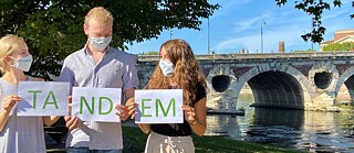 Deux jeunes femmes et un jeune homme avec des masques et les lettres TANDEM sur une prairie devant un pont