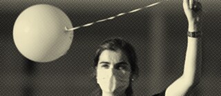 Mια κοπέλα με καστανά μαλλιά έχει σηκωμένο το αριστερό της χέρι, κρατώντας ένα άσπρο μπαλόνι από ένα κορδόνι. Φορά μαύρο μπλουζάκι και άσπρη προστατευτική μάσκα στο στόμα και τη μύτη.