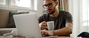 Ein junger Mann sitzt entspannt mit einer Kaffeetasse zu Hause am Laptop und arbeitet.