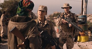 Zwei Bundeswehrsoldat*innen, ein Mann und eine Frau, Maschinengewehr im Arm, sprechen mit einer kurdischen Kämpferin