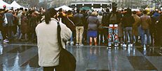A group of people seen from behind in a rainy city; in the foreground a person pointing a camera at the group of people.