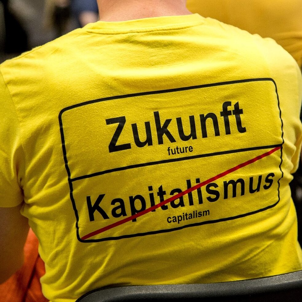 Eine an einer Tagung teilnehmende Person trägt ein T-Shirt mit der Aufschrift "Zukunft | Kapitalismus (durchgestrichen) " im Stile eines Ortsausfahrtschildes zeigt.