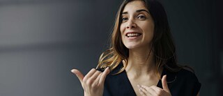 Porträt einer redenden, gestikulierenden Frau vor dunklem Hintergrund.