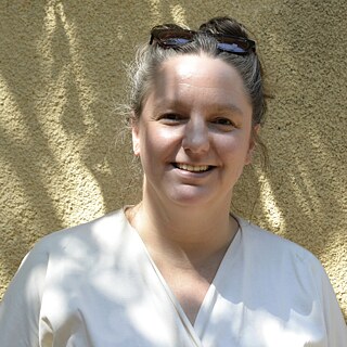 The picture shows a portrait of a woman (Ulrike Syha). She has tied back her hair and is wearing a white top and pushed up sunglasses. She is standing in front of a wall.