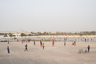 Ein unbebautes Stück Land wird zu improvisierten Sportplätzen umfunktioniert. Im Vordergrund spielen hauptsächlich aus der afrikanischen Diaspora stammende Männer Fußball, während Mitglieder der südasiatischen Gemeinschaft auf dem Asphalt des leeren Parkplatzes Kricket spielen.