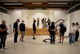 Twelve people are seen looking at or trying out the artworks. One of them is an installation made of tubes, the other one is a sculpture made of strings, a bow and a riding helmet.