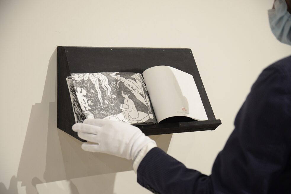 A hand in a white cloth glove is seen leafing through a book of artworks. 