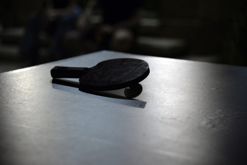 The image shows a table tennis ball lying on a table tennis table in the dark. On the ball lies a table tennis bat.