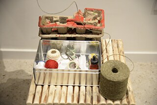 The image shows a stack of egg cartons and in front of it a metal box with thread and bandages. The whole thing is on a wooden base.