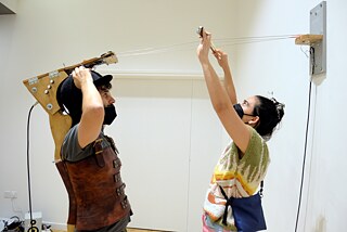 You can see two people from the side. One of the persons is wearing a construction made of wood, with a riding helmet on his head and a leather harness.  Strings go from the construction towards the wall. The second person is playing on the strings with a bow.
