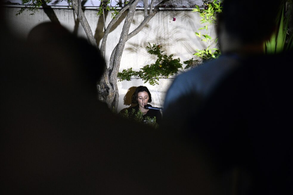 Das Bild zeigt eine Frau, die draußen vor einer Wand sitzt. Links und rechts von ihr sieht man Pflanzen. Sie hat den Mund zum Sprechen geöffnet und etwas unterhalb ihres Gesichts befindet sich ein Mikrophon.