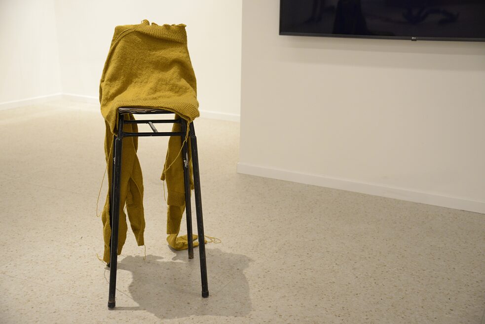 The image shows a chair. On the chair is a mustard yellow sweater. This one has several arms. There are longer threads hanging from the end of the sweater.