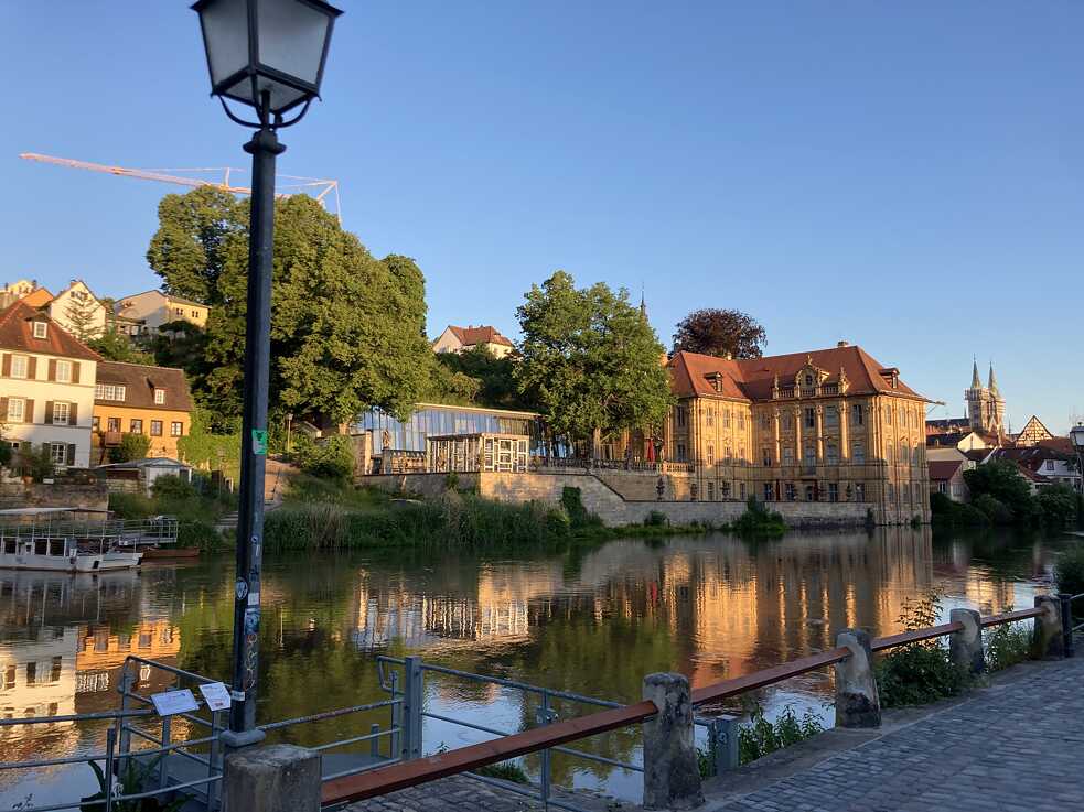 Ein Fluss, im Vordergrund ein gepflastertes Ufer und eine Laterne, am linken Bildrand an der anderen Uferseite eine Fähre oberhalb des Ufers mehrere Gebäude und Bäume