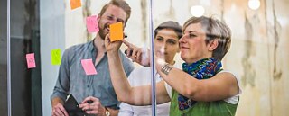 Drei Teamkollegen arbeiten kreativ mit Post-its an einer Glaspinnwand. Sie sind sehr konzentriert und haben Spaß.