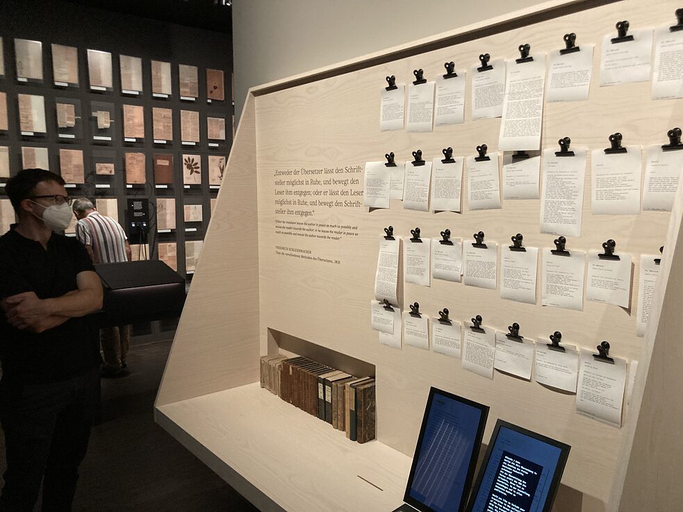 A museum wall on which printed slips of paper are hung in several rows with black staples.