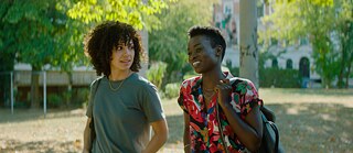 Zwei junge Frauen unterhalten sich, eine von ihnen lacht. Im Hintergrund sieht man von der Sonne angestrahlte Bäume und einen Platz.