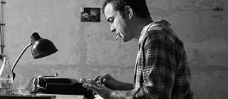 Ein Mann sitzt, mit einer Zigarette im Mundwinkel, an einem Schreibtisch und tippt auf seiner Schreibmaschine