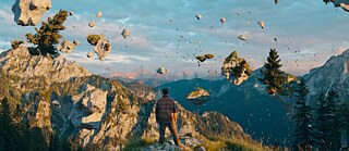 Ein Mann steht hoch oben an einem Aussichtspunkt im Gebirge und blickt ins Tal. Zu sehen ist er von hinten. Um ihn herum scheinen Gesteinsbrocken und Bäume zu schweben.