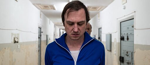 Ein Mann in blauer Jacke wird von einem Wärter durch einen Gefängnistrakt geführt. Sein Blick ist nach unten gerichtet.