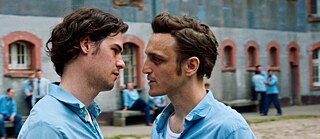 Zwei Männer in blauen Hemden schauen sich in die Augen. Hinter ihnen sitzen mehrere Männer in der gleichen Montur vor einem Gebäude herum.