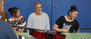 Zwei Mädchen sitzen lachend an einem Tisch auf dem eine Gitarre liegt. In ihrer Mitte sitzt ein lächelnder, mittelalter Mann.