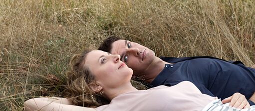 Eine Frau und ein Mann mit auffällig blauen Augen liegen auf einem Feld. Die Frau blickt in den Himmel während der Mann sie anschaut.