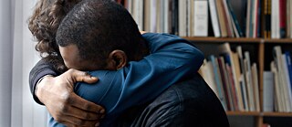 Zwei Menschen umarmen sich fest. Im Hintergrund ist ein Regal voller Bücher zu erkennen. 