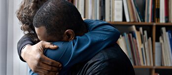 Zwei Menschen umarmen sich fest. Im Hintergrund ist ein Regal voller Bücher zu erkennen. 