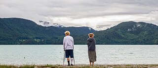 Ein junger, sich auf Krücken stützender Mann und eine Frau stehen mit Abstand zueinander vor einem See und blicken voneinander weg. 