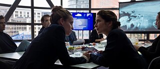 Zwei Frauen sitzen in ein enges Gespräch vertieft an einem Tisch. Eine von ihnen hält einen Stapel Fotos in den Händen. Um sie herum sitzen Männer die sie mustern.