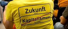 Eine an einer Tagung teilnehmende Person trägt ein T-Shirt mit der Aufschrift "Zukunft | Kapitalismus (durchgestrichen) " im Stile eines Ortsausfahrtschildes zeigt.
