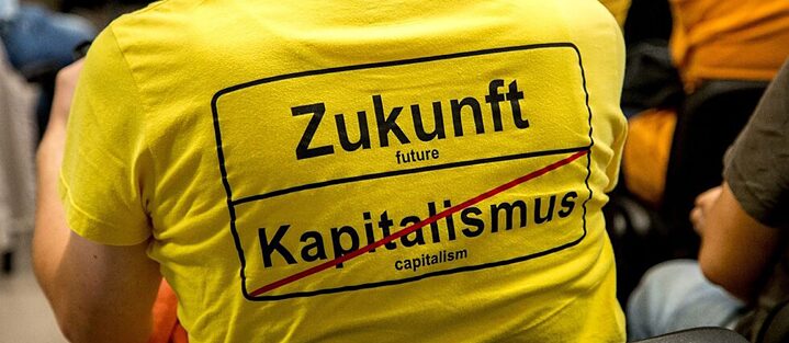 A person attending a meeting wears a T-shirt with the inscription "Future | Capitalism (crossed out) " in the style of a town exit sign shows.