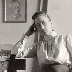 The image is in black and white. You can see a woman (Hilma af Klint) sitting on a chair and leaning against a table on the left. Behind her in the room is a portrait.