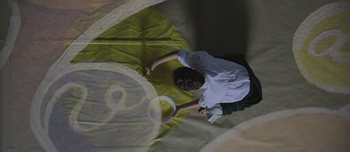Η φωτογραφία δείχνει μια γυναίκα (Hilma af Klint), που ζωγραφίζει ξυπόλητη γονατιστή στο πάτωμα. Το έργο τέχνης είναι άσπρο, πράσινο και ανοικτό μωβ.