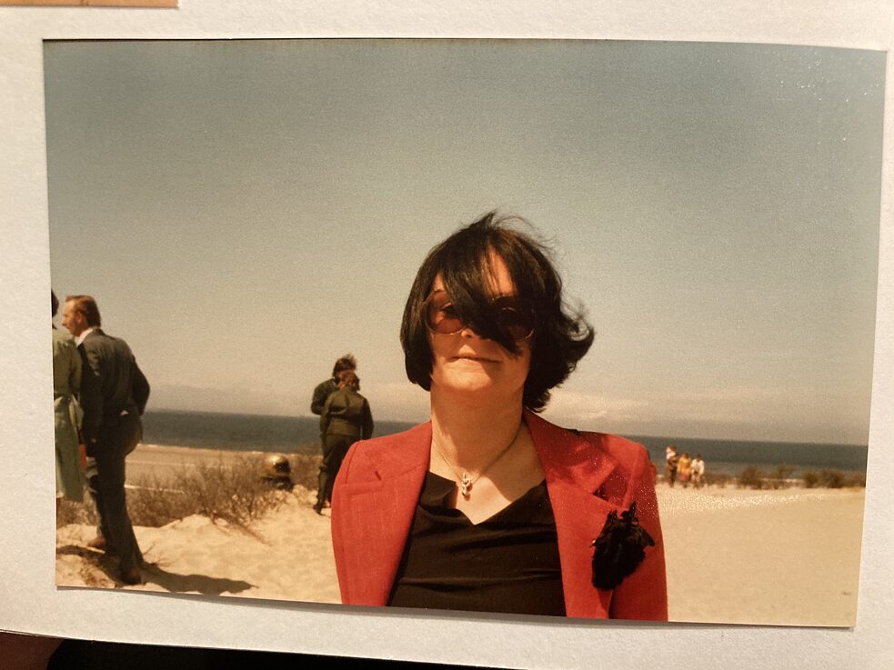 Eine Fotografie, auf einer Unterlage liegend, zeigt eine Frau mit getönter Brille, eine Haarsträne verdeckt einen Teil ihres Gesichts; im Hintergrund Menschen, Strand, Meer.