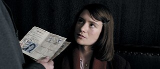 Sophie Scholl (Julia Jentsch) auf der rechten Seite, den Blick nach oben gerichtet. Unten links kann man ihren Reisepass sehen.