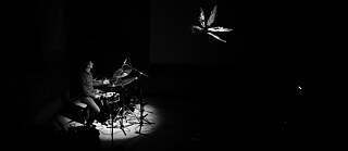 The black and white photo is mainly dark. In the left side of the picture, a drummer is illuminated, sitting in front of his drum kit. His face is turned away from the camera. In the upper right part of the picture you can see the contours of a butterfly.