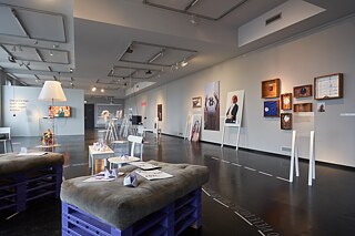 Ein heller Ausstellungsraum mit einigen Bildern und Gemälden an der Wand und Sitzplätzen sowie Aufsteller im Raum verteilt.